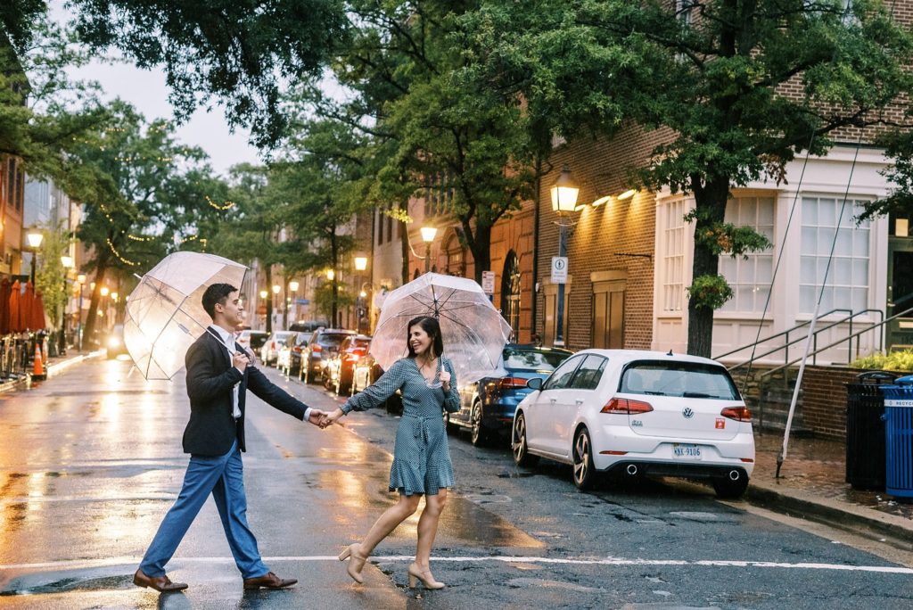 engaged couple walks under umbrellas through crosswalk in Old Town Alexandria